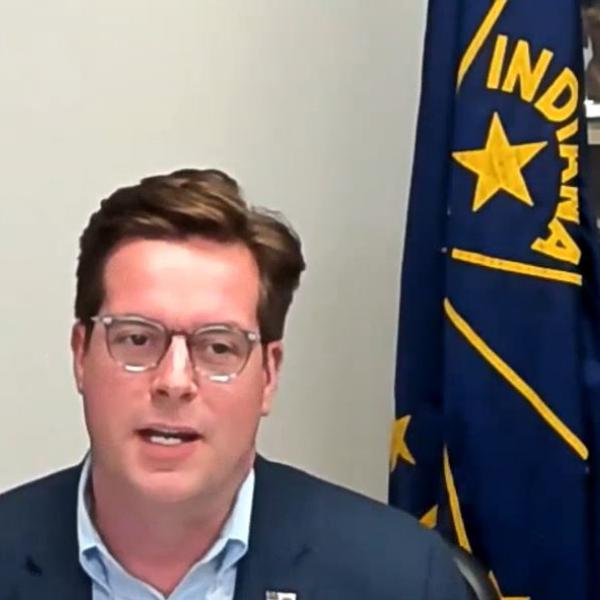 A white man with short brown hair and glasses sits next to and in front of an upstanding flag of Indiana.