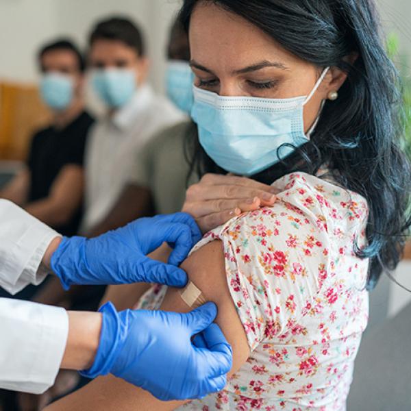 Doctor wearing blue latex gloves placing a bandage on the shoulder of a woman with long black hair who is wearing a mask.