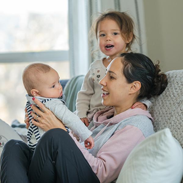 Young mother with brown pulled back hair holds a baby on a light gray couch while a toddler looks on