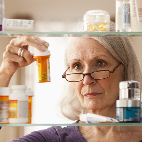 An older white woman with shoulder length gray hair wears brown glasses while looking at a vial of pills in front of a medicine cabinet