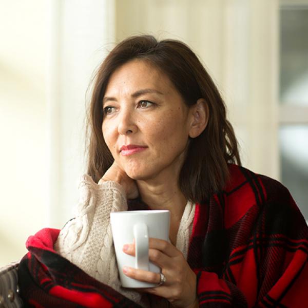 White woman with shoulder length brown hair with red plaid blanket around shoulders holds mug and looks out window