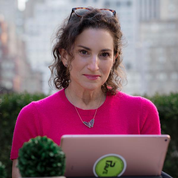 White woman with shoulder length brown curly hair wearing a pink sweater and butterfly necklace with open laptop in front of a city skyline