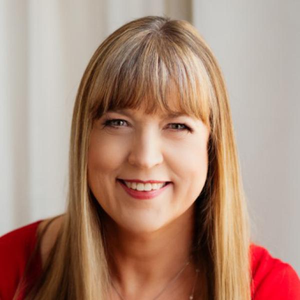 White woman with long brown hair and short bangs smiles and wears a red blouse