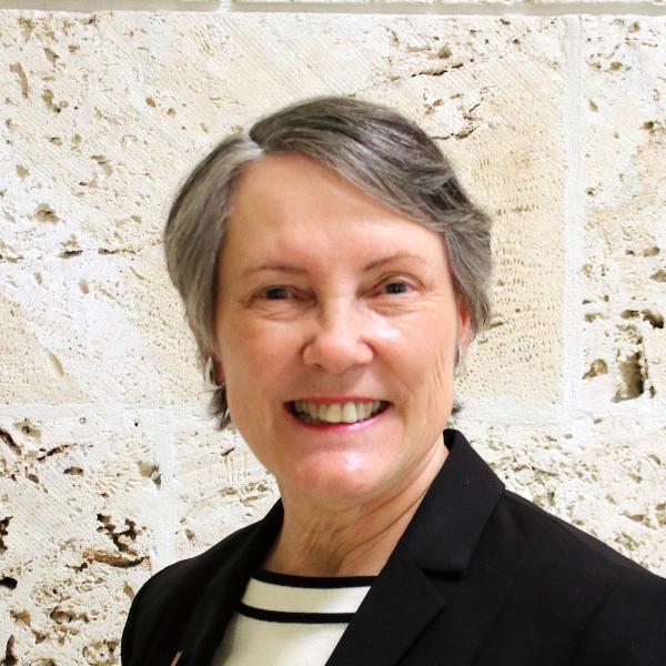 A smiling white woman with short gray sandy hair stands in front of a cream stone wall wearing a black blazer.