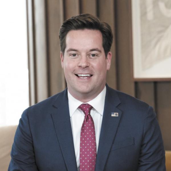 White man with short thick brown hair and wearing a blue jacket over a white button down shirt with maroon tie smiles at the camera 