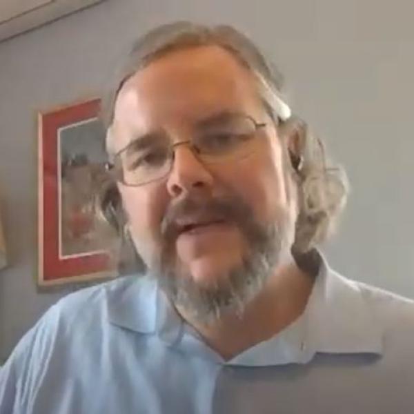 A white man with a beard glasses and shoulder length hair wearing a light blue polo shirt sits in front of a light blue wall with several framed pictures