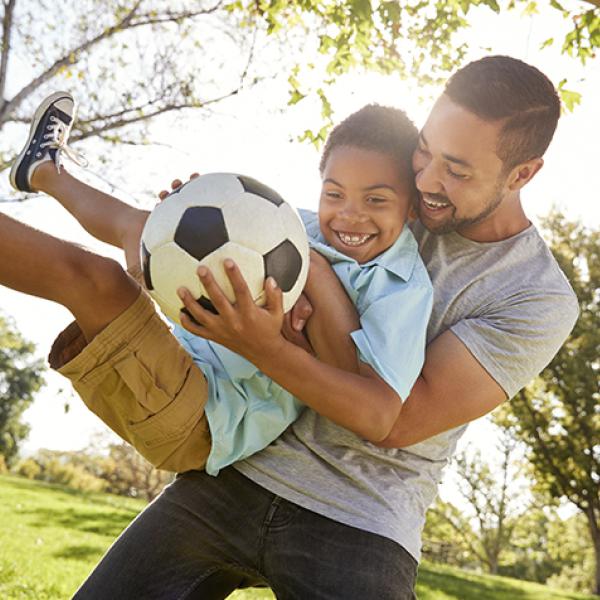 Young black man in gray T-shirt  lifts African American boy who is holding a soccer ball and wearing a light blue polo shirt and light brown cargo shorts