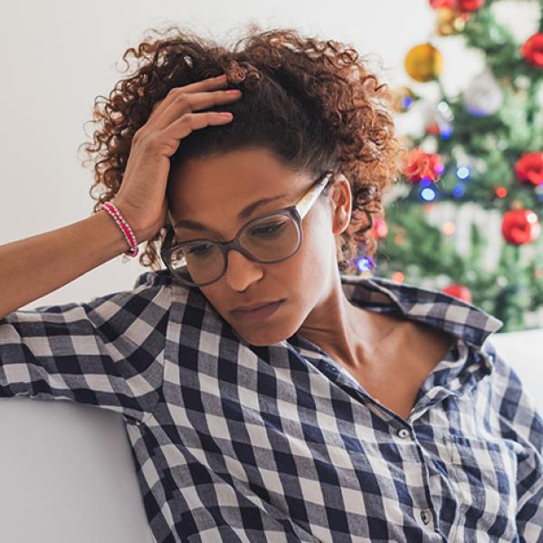 Black woman wearing glasses and wearing a black and white button down shirt looks dejected while sitting in front of a Christmas tree
