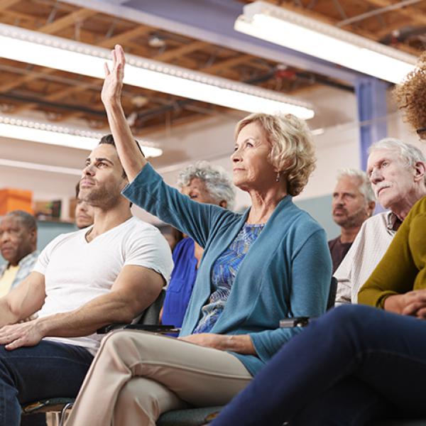 An older white woman with short blonde hair wearing a teal sweater over a blue patterned blouse raises her hand at a meeting 