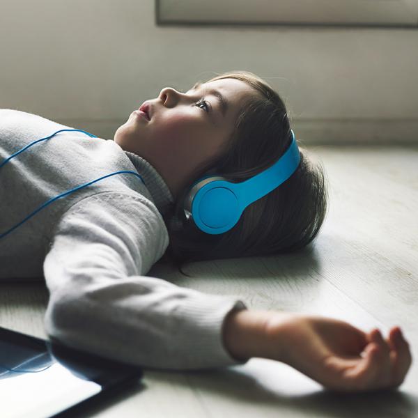 A young white boy with brown hair wearing a gray sweater lies on a gray wooden plank floor and wears light blue headphones