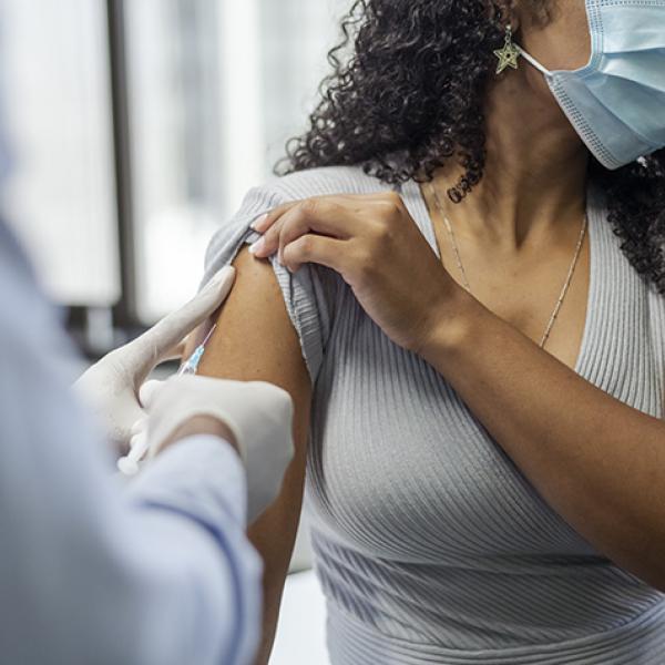 A doctor wearing latex gloves inserts a syringe into the upper right arm of a woman who has curly black shoulder length hair and is wearing a mask.