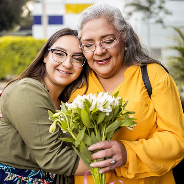 Daughter with long brown hair wearing glasses hugs mother, who has long gray hair and glasses and is holding flowers