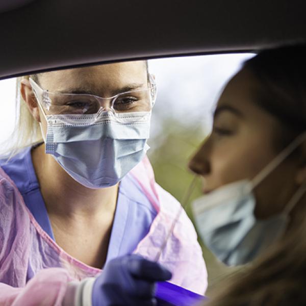 Woman wearing mask and safety glasses takes a nasal swab from a woman through an open car window.