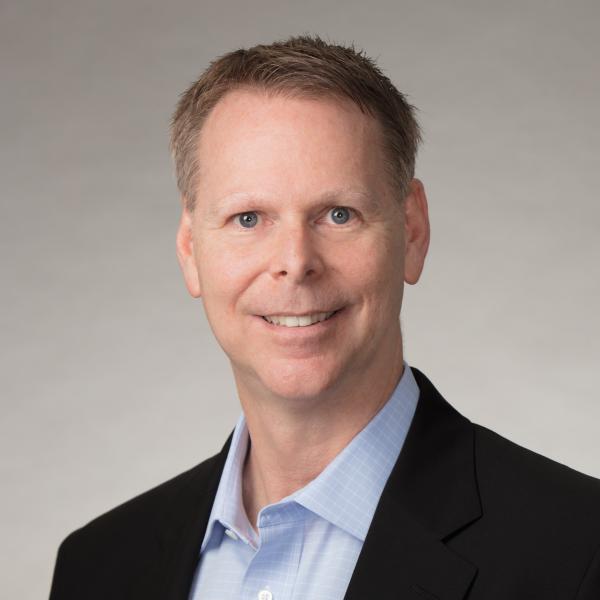 Middle aged white man with short hair wearing a black blazer over a light blue shirt.