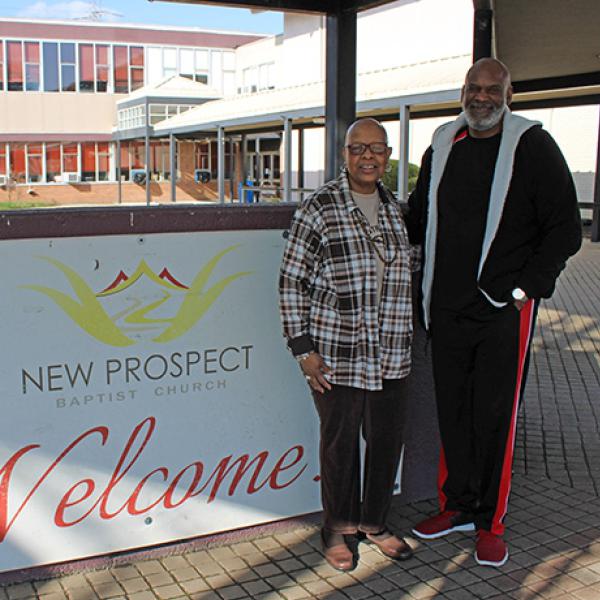 A bald Black woman  wearing glasses and a plaid shirt and a tall bald Black man in a jacket and sweatpants stand outside in front of a sign for New Prospect Baptist Church.