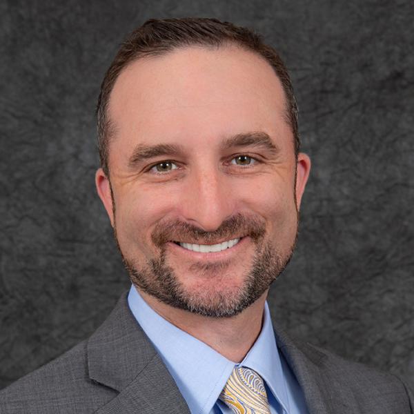 Middle aged white man with brown hair and eyes and a beard wearing a gray suit with a light blue shirt and yellow tie