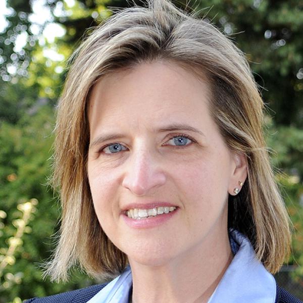 White woman with blue eyes and shoulder length straight blonde hair smiles at the camera in an outdoor setting