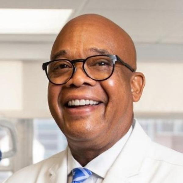 Smiling bald middle aged Black man wearing round black glasses and a white lab coat.